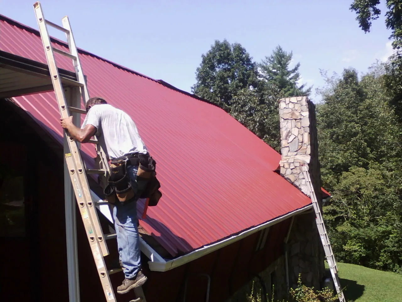 Roofer on a ladder beside a red metal roof and stone chimney — Soffit & Fascia Repair by Roofing Brothers in Steger